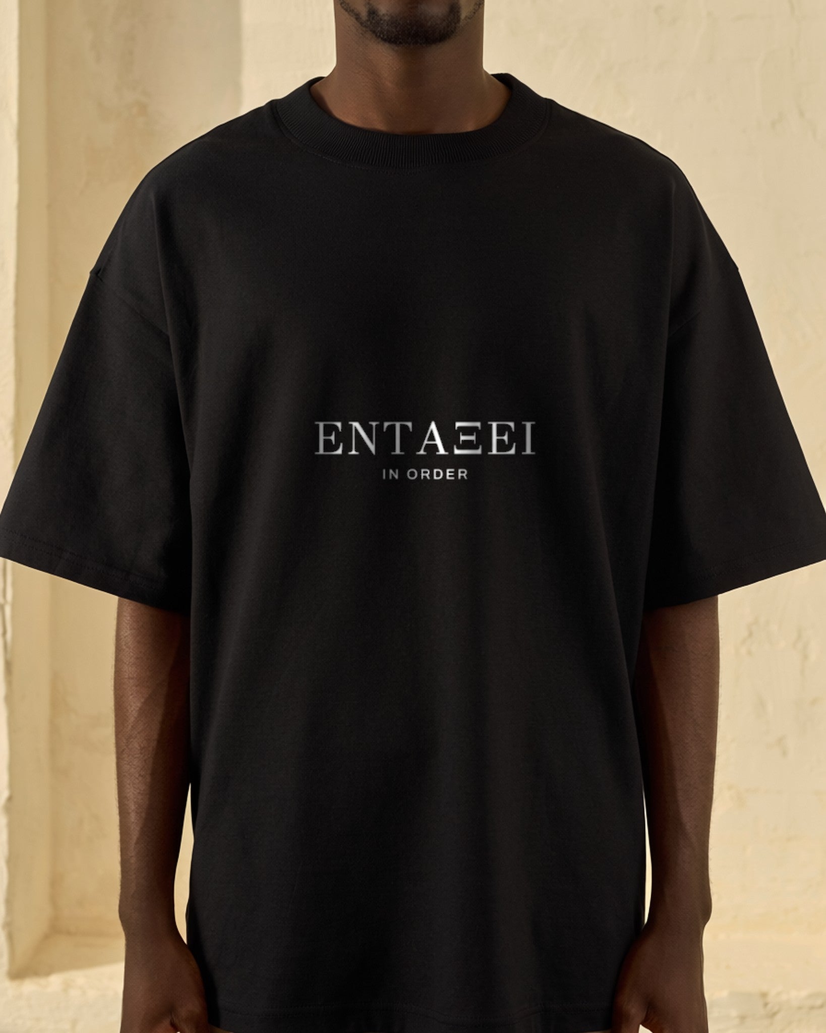 Model wearing the black ΕΝΤΑΞΕΙ House Tee against a sunlit cream plaster wall, ΕΝΤΑΞΕΙ printed across the chest with the gloss IN ORDER beneath.