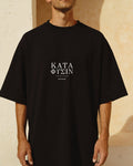 Model wearing the black According to Nature Tee against a sunlit cream plaster wall, ΚΑΤΑ ΦΥΣΙΝ printed across the chest in Greek Didot capitals.