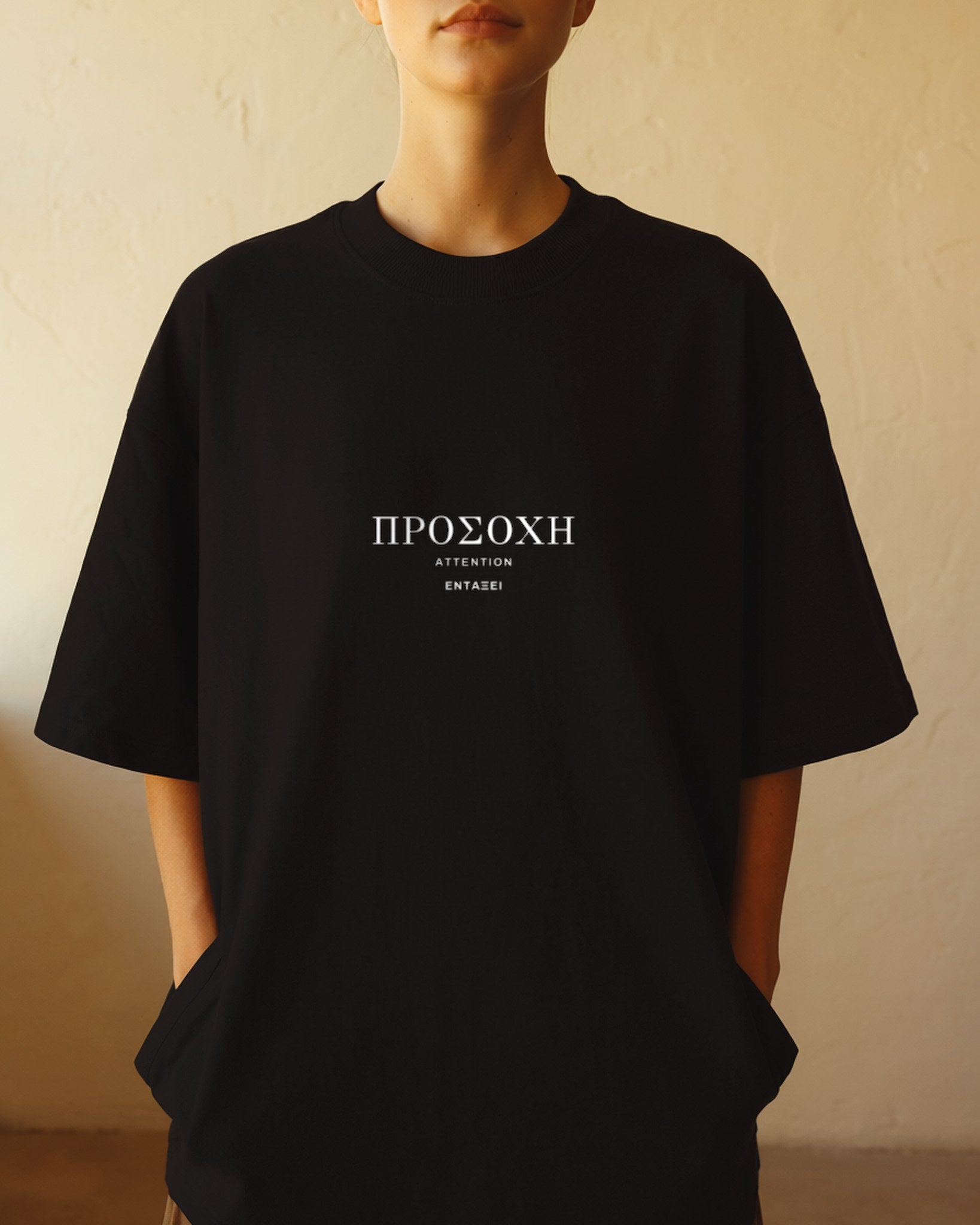 Model wearing the black Attention Tee against a sunlit cream plaster wall, ΠΡΟΣΟΧΗ printed across the chest in Greek Didot capitals.