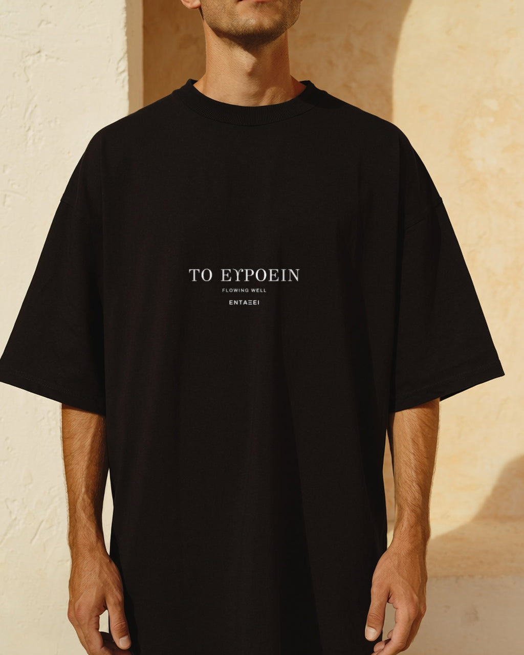 Model wearing the black Flowing Well Tee against a sunlit cream plaster wall, ΤΟ ΕΥΡΟΕΙΝ printed across the chest in Greek Didot capitals.