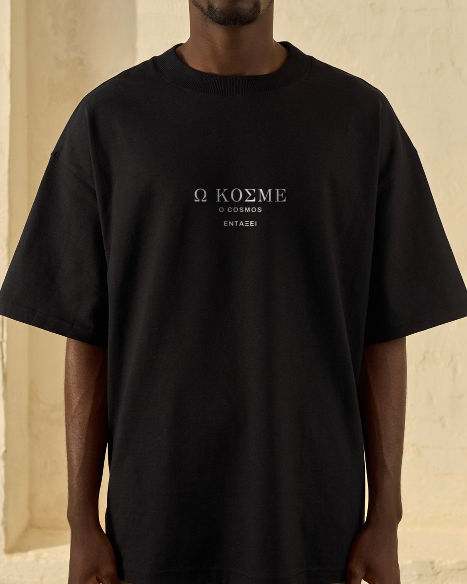 Model wearing the black O Cosmos Tee against a sunlit cream plaster wall, Ω ΚΟΣΜΕ printed small at the centre chest in Greek Didot capitals.