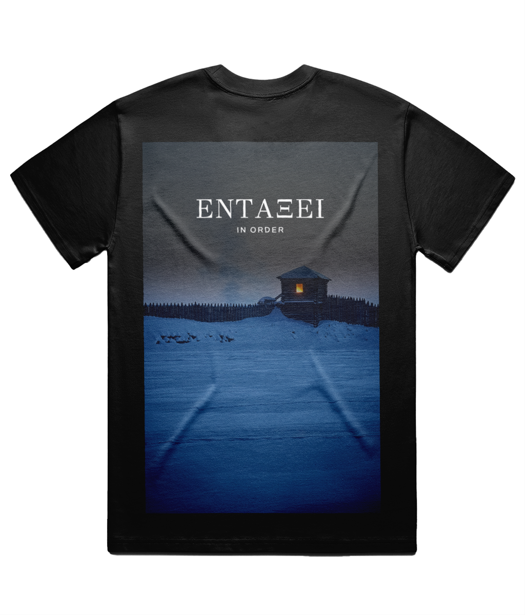 Black Carnuntum Tee shown flat on white from the back, full back print: a night photograph of the frozen Danube camp, one lit window in a timber watchtower, with ΕΝΤΑΞΕΙ and IN ORDER set above it in Didot.